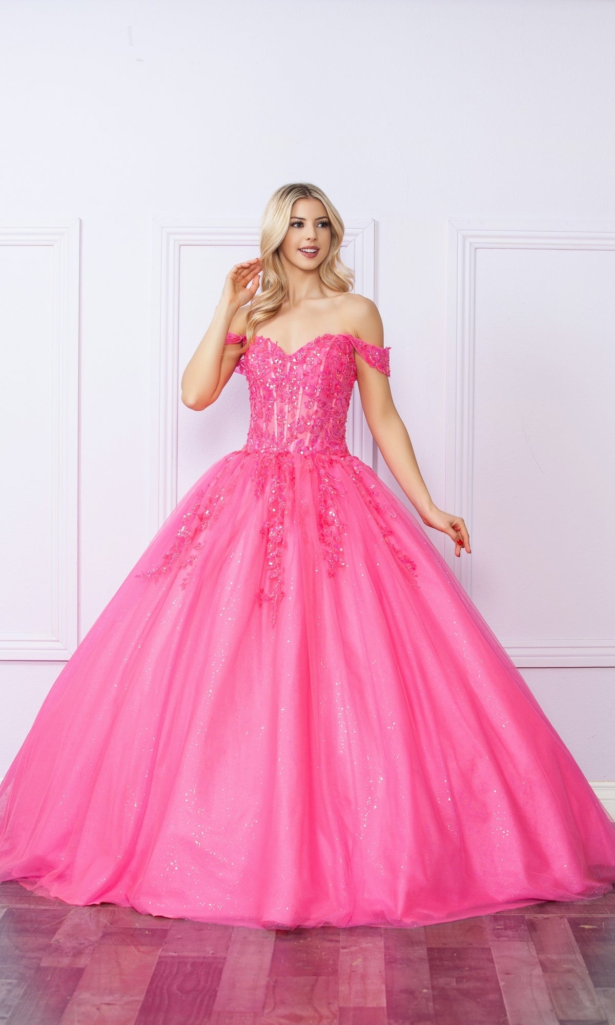 A woman in the Bright Off-the-Shoulder Long Prom Ball Gown H1349, featuring a vibrant pink hue, full skirt, and sequin details, stands against a white paneled wall.